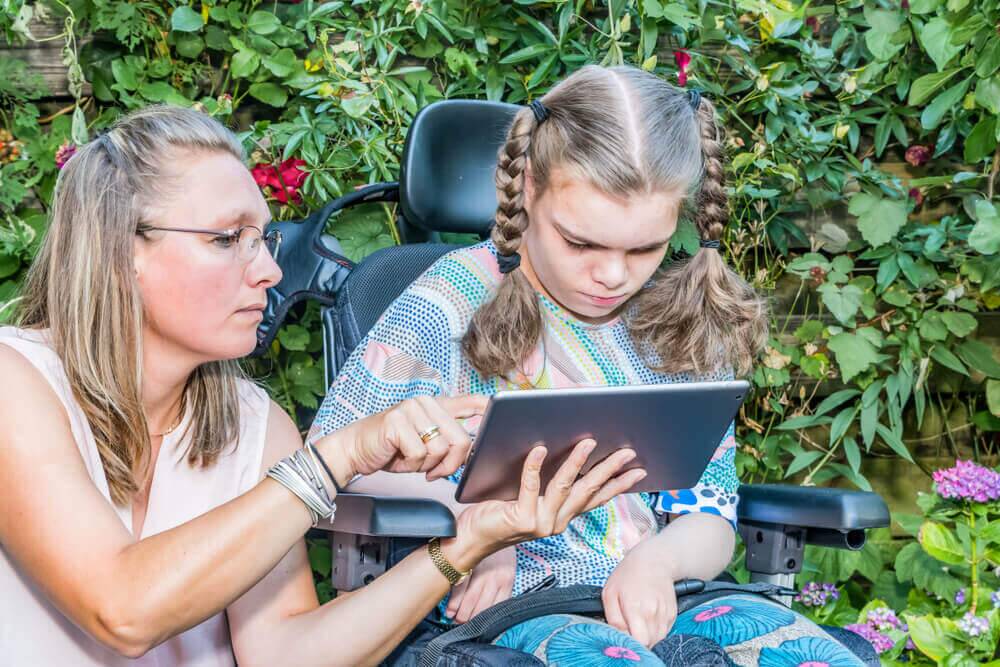 Disability a disabled child in a wheelchair sitting outside looking at a tablet computer together with a voluntary care worker