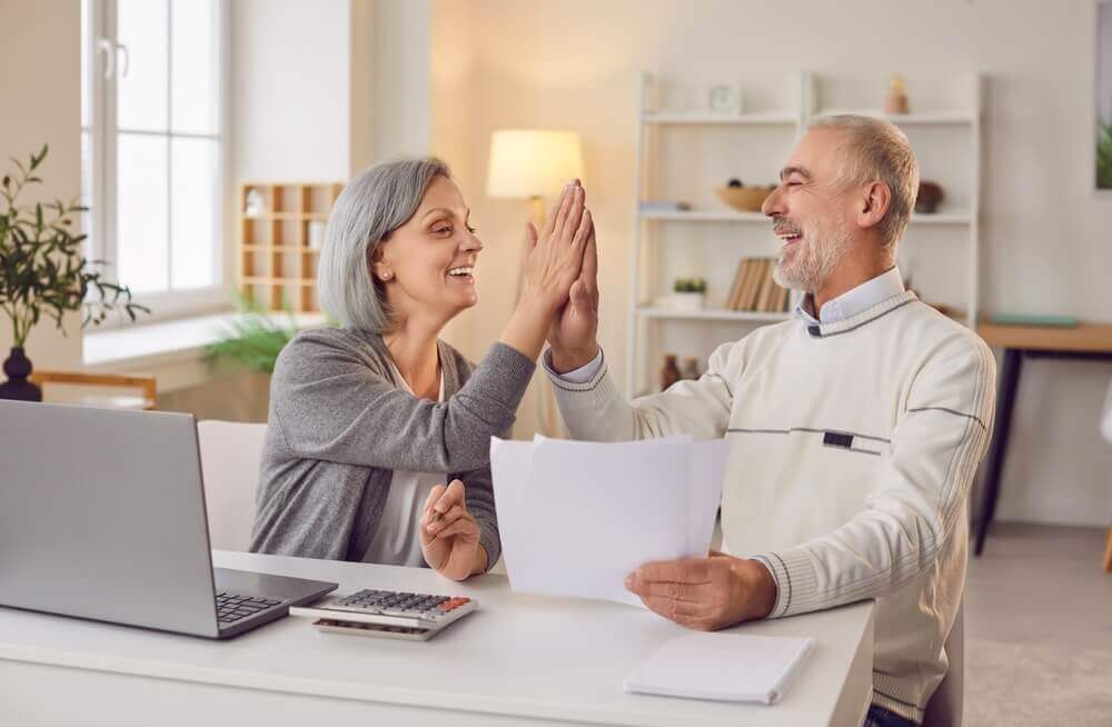 Senior couple sitting at the table with laptop and bills giving high five each other calculating finances or taxes at home. Elderly retired man and woman rejoicing income and profit on pension.